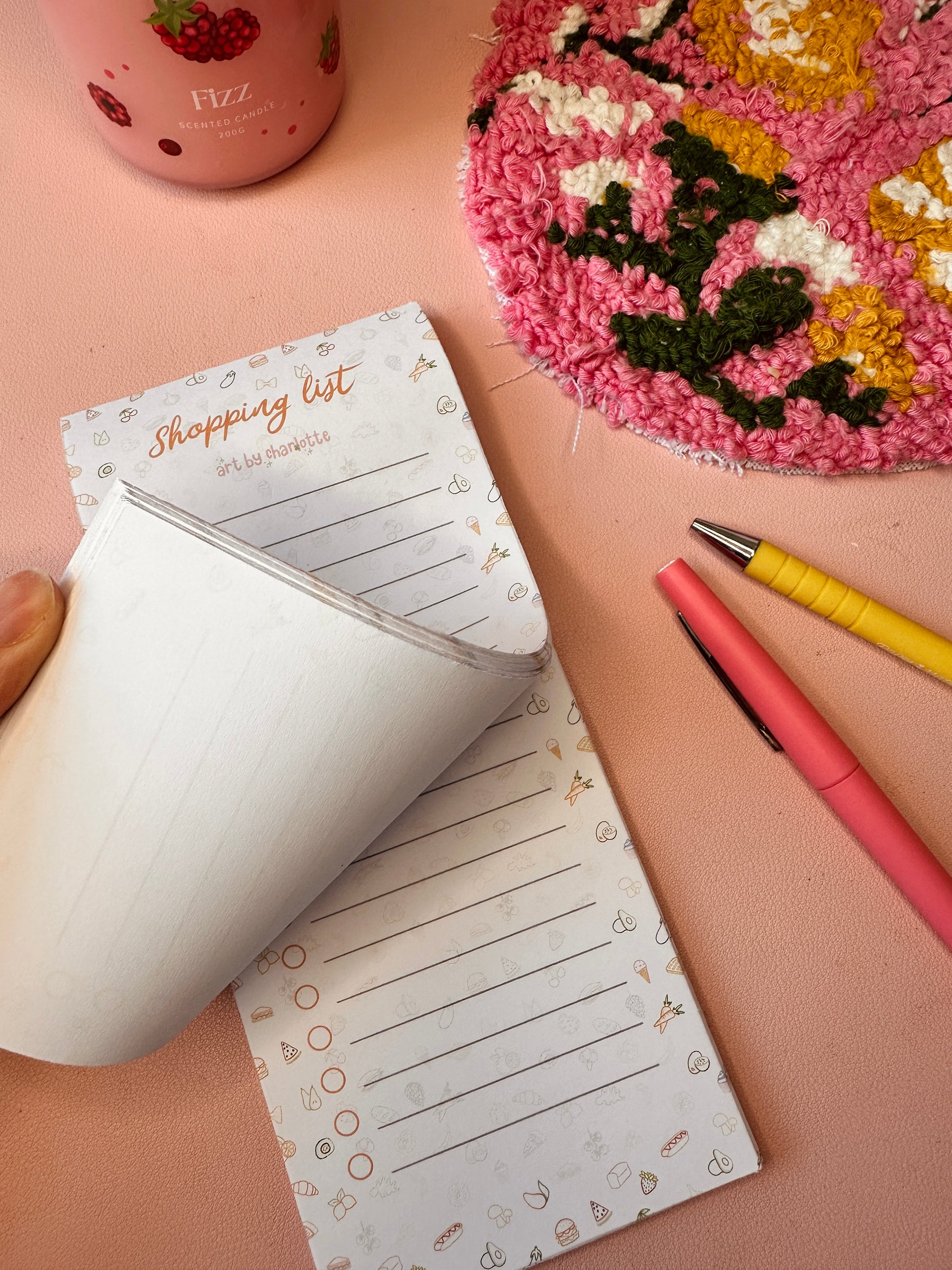 Notepad with a shopping list on a pink surface with a decorative item and pens.