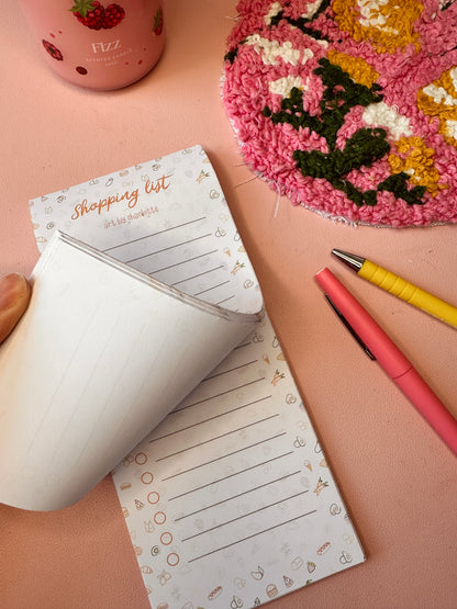Notepad with a shopping list on a pink surface with a decorative item and pens.