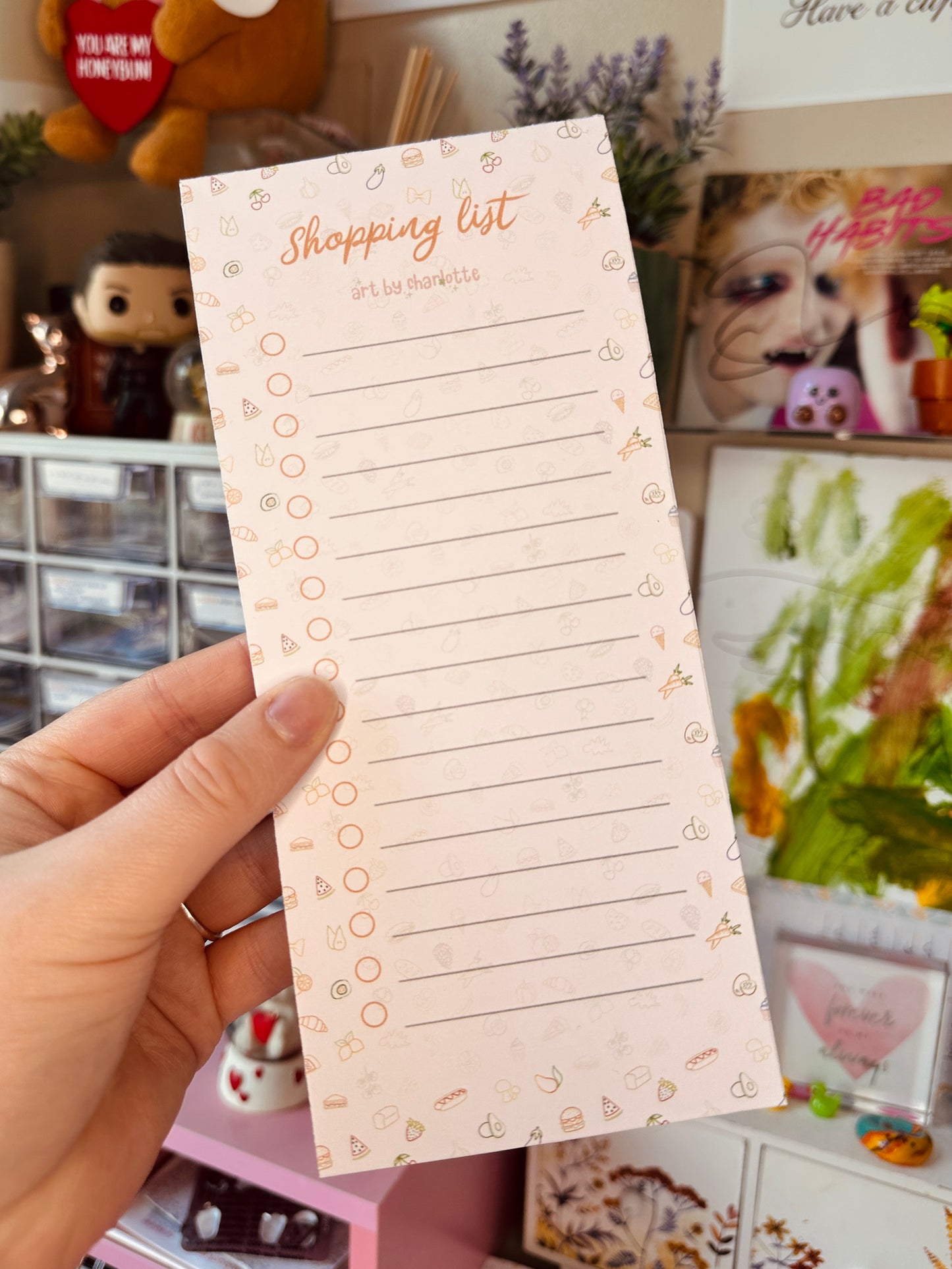Hand holding a shopping list pad with decorative elements against a cluttered background