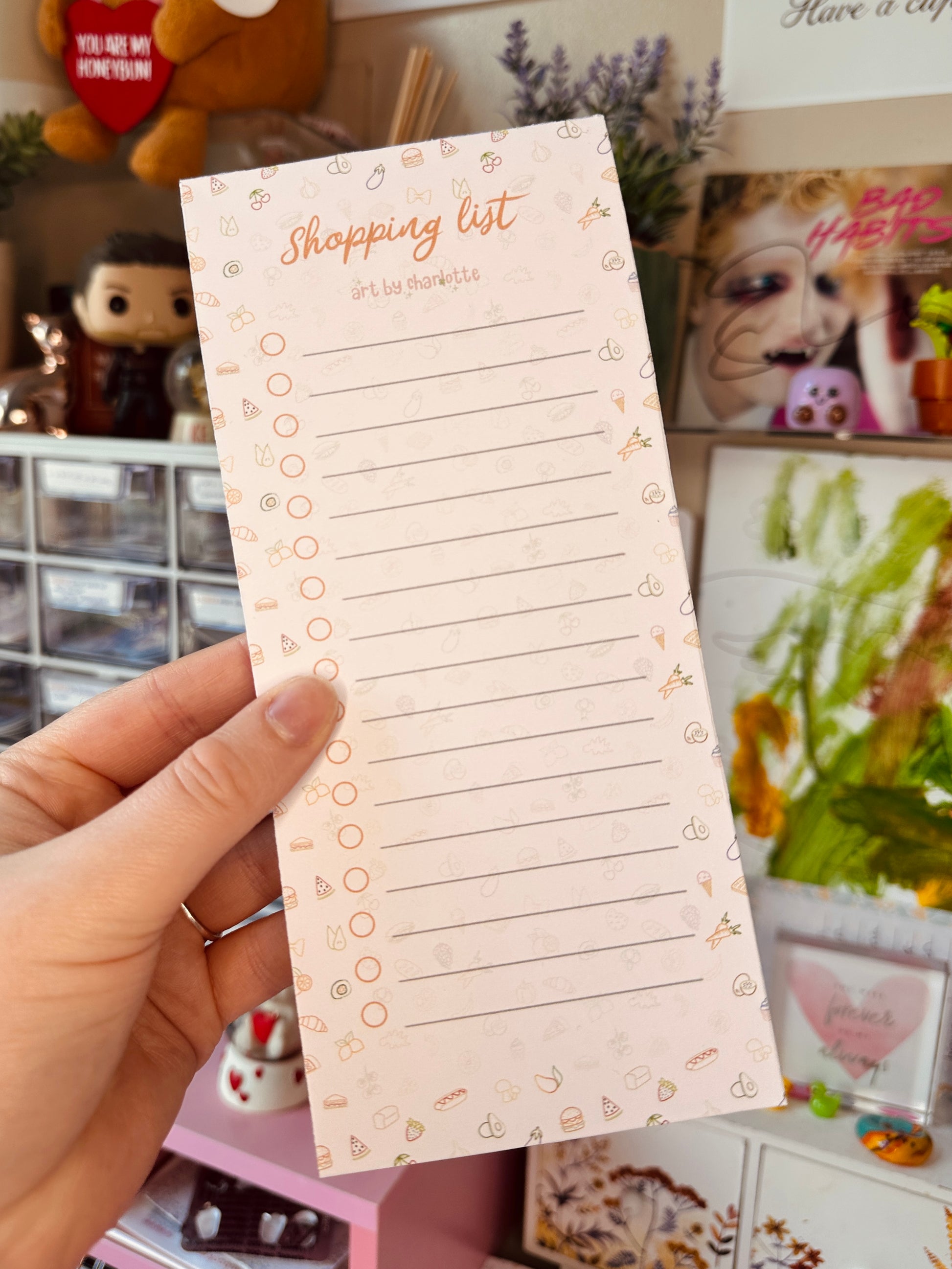 Hand holding a shopping list pad with decorative elements against a cluttered background