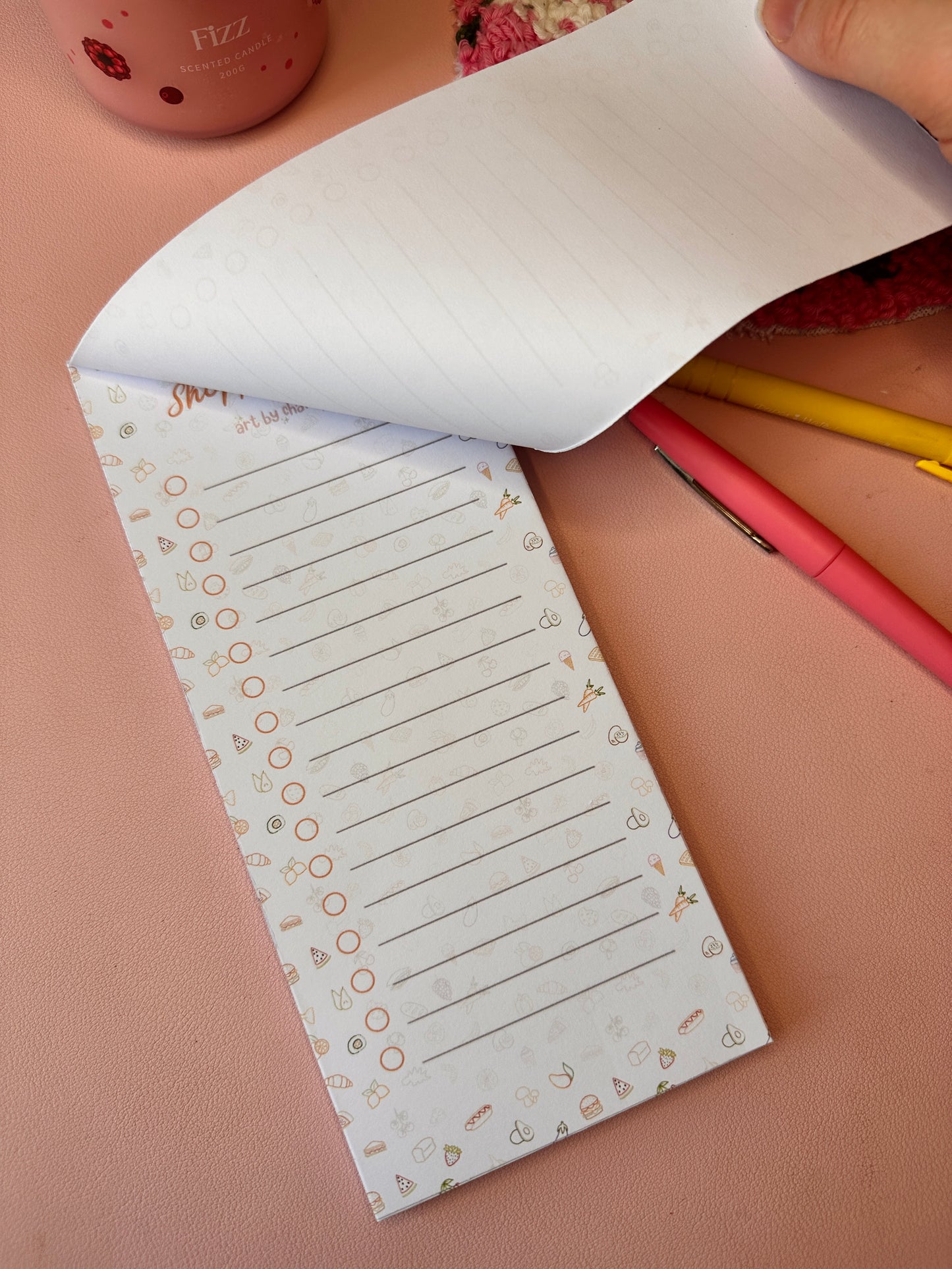 Notebook with lined pages and decorative border on a pink surface with pens.