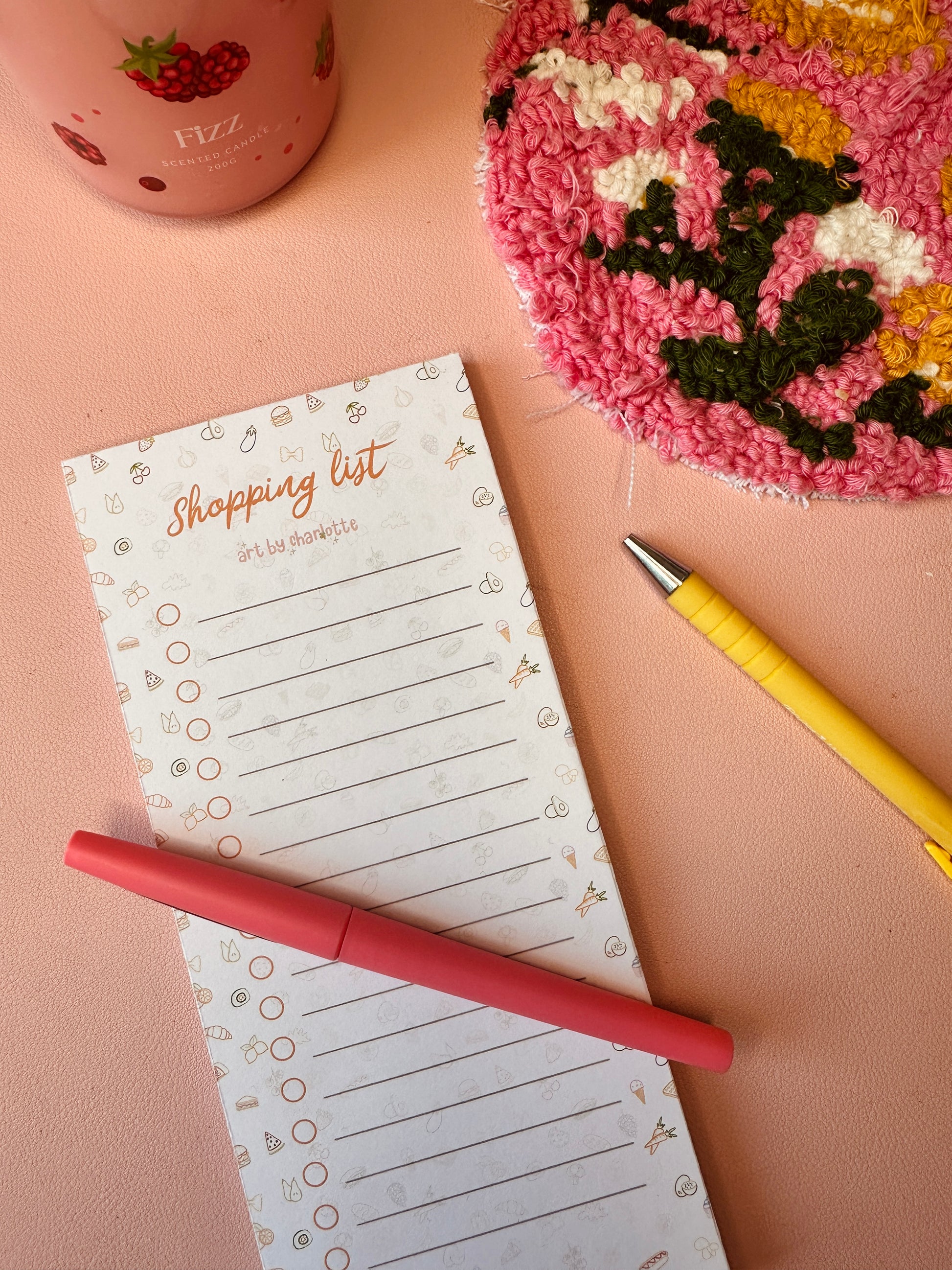 Notepad with 'Shopping list' on a pink surface with a pink pen and colorful fabric.